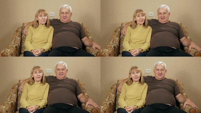 老年夫妇在家躺在沙发上休息，看着相机。他们在微笑。丈夫拥抱妻子