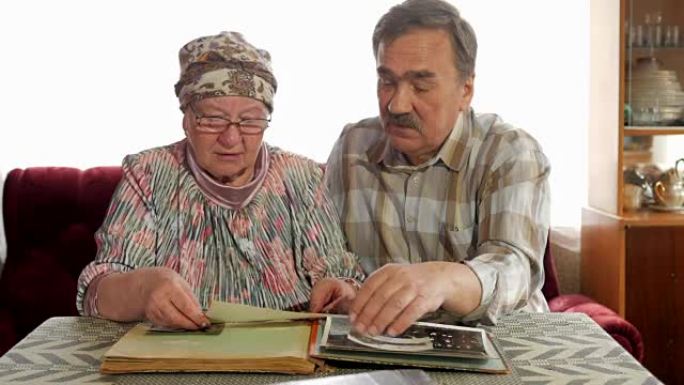 一对老年夫妇正在家里看着他们的旧照片，聊天。有胡子的男人，有眼镜的妻子