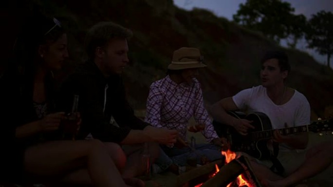 晚上，一群年轻开朗的人坐在海边的火炉旁，烤香肠和弹吉他。慢动作镜头