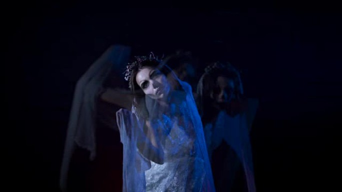 穿着白色婚纱和面纱的年轻新娘的幻影移动了她的手。万圣节化妆的恶作剧女孩站在黑色背景下。恐怖中的神秘女