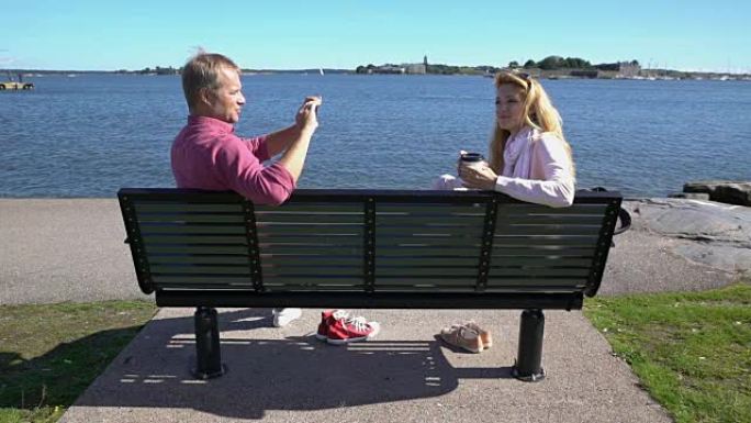 一对在海边的长凳上。年轻人正在用智能手机给她的女朋友拍照。慢动作。