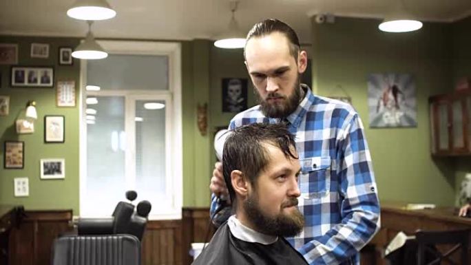 迷人的年轻理发师正在用剪刀剪头发。他专注地看着头发。大胡子的人抬起下巴