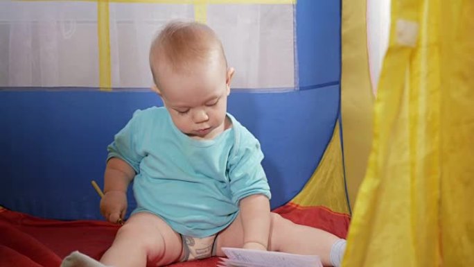 迷人的婴儿在房子里的儿童五颜六色的帐篷里玩耍。这个男孩小心翼翼地在铅笔笔记本上画画