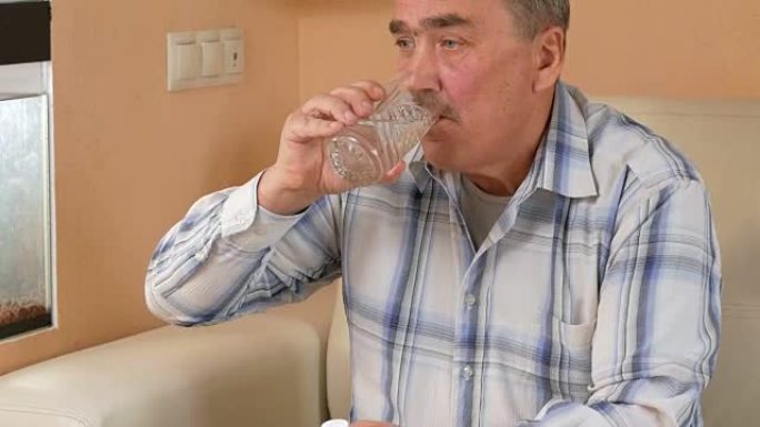 老人胡子在家吃药，喝一杯水。他坐在水族馆附近的沙发上，改善了他们的健康