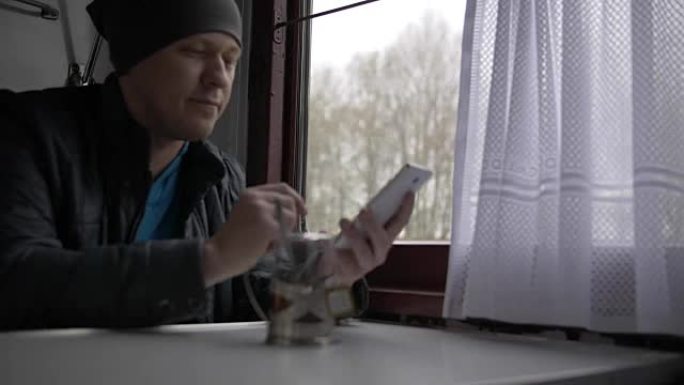 一个年轻人正戴着灰色的帽子和灰色的外套，坐在桌子旁，用白色智能手机浏览信息。窗外闪烁着树木