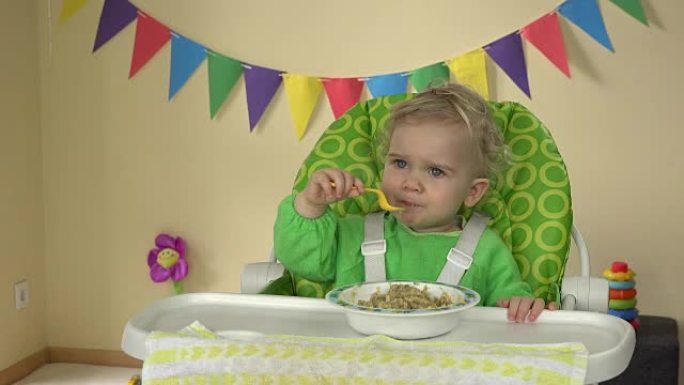 饥饿的勺子女婴坐在喂食椅上，从盘子里吃粥
