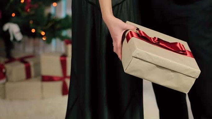 穿着黑色晚礼服的女人手里拿着一条红丝带的礼物，一个穿着黑色西装的男人在一个有壁炉和圣诞树的白色房间里