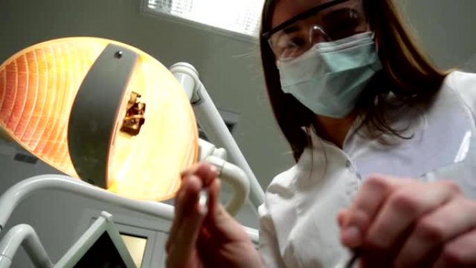 戴着口罩的年轻女牙医带着工具走近，拿着口腔镜和牙医的钩子，牙科灯灯进入病人的嘴里，站在病人身上，看着