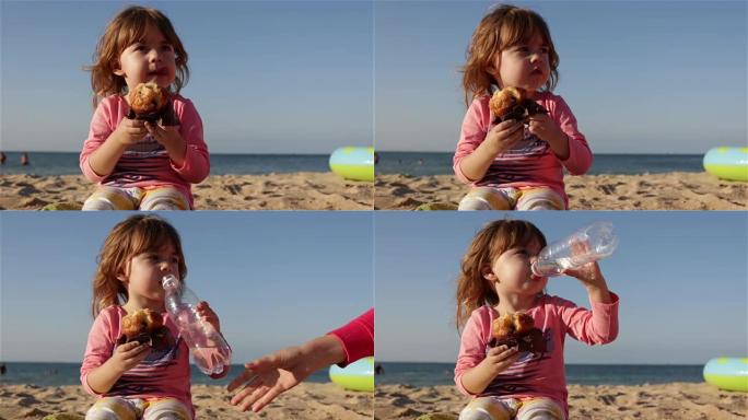 一个可爱的小孩吃纸杯蛋糕，从瓶子里喝水。她坐在海边的海滩上