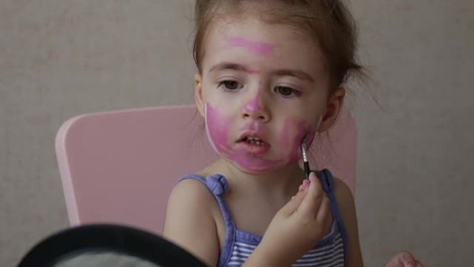 金发三岁的婴儿看着镜子在脸上画彩色条纹