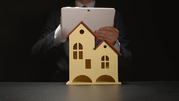 商人拿着平板电脑放在桌子上的房屋模型后面