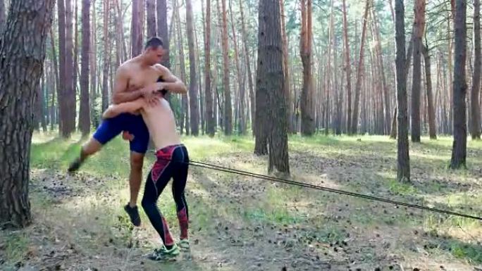 年轻的运动男子裸露，裸露的躯干，练习打击，捕捉，战斗的技术，用橡皮绳，慢动作。在松树林中，在夏天，在