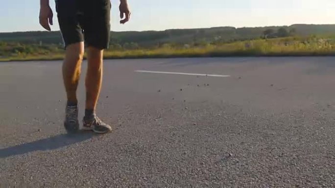 男性的脚在日落时在乡村道路上行走，靠近。年轻人正走在一条空旷的乡间小路上。穿过阳光照射的人。小男孩的