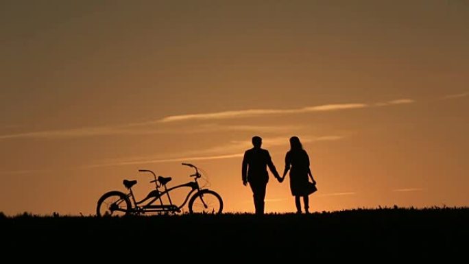 美丽夫妇的剪影与双人自行车手牵着手对抗神话般的日落背景