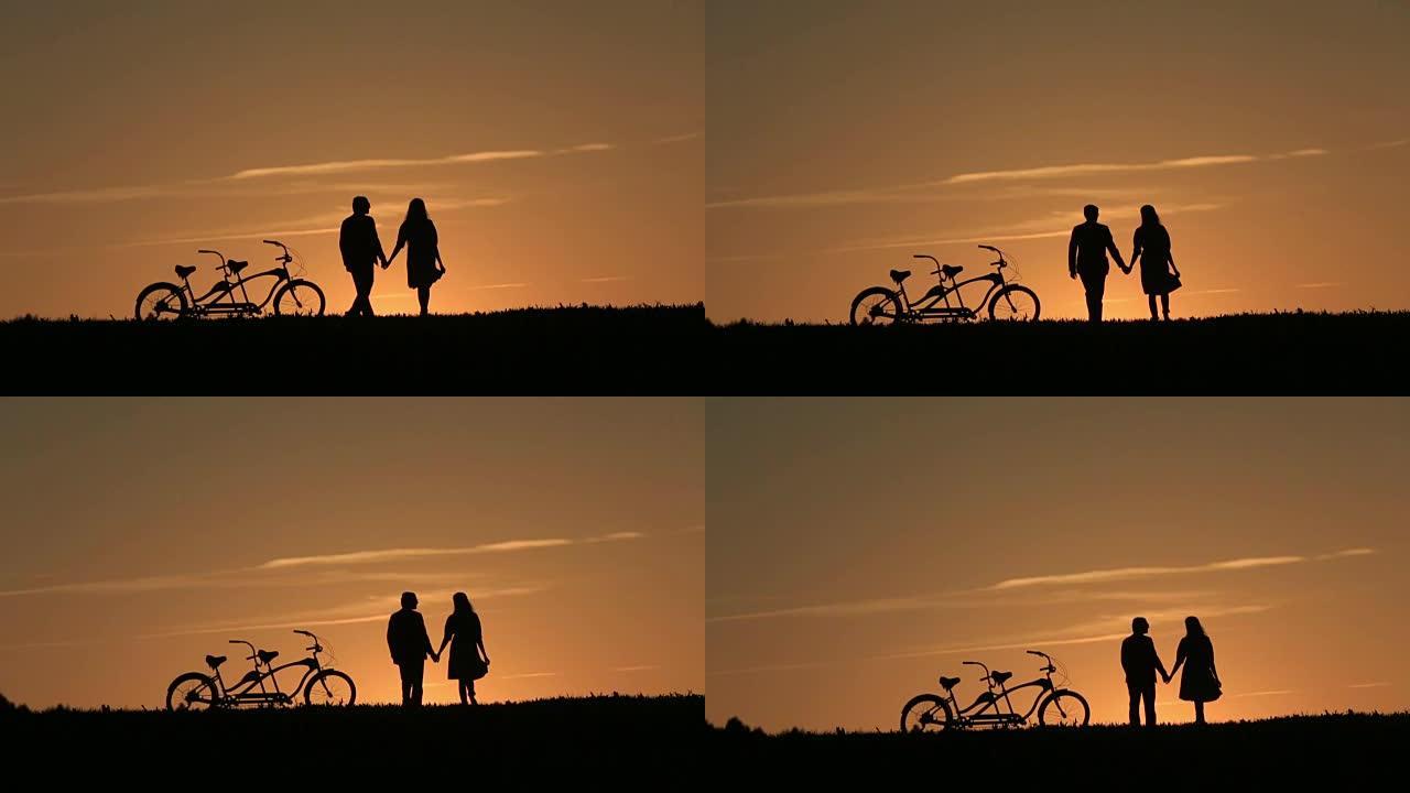 美丽夫妇的剪影与双人自行车手牵着手对抗神话般的日落背景