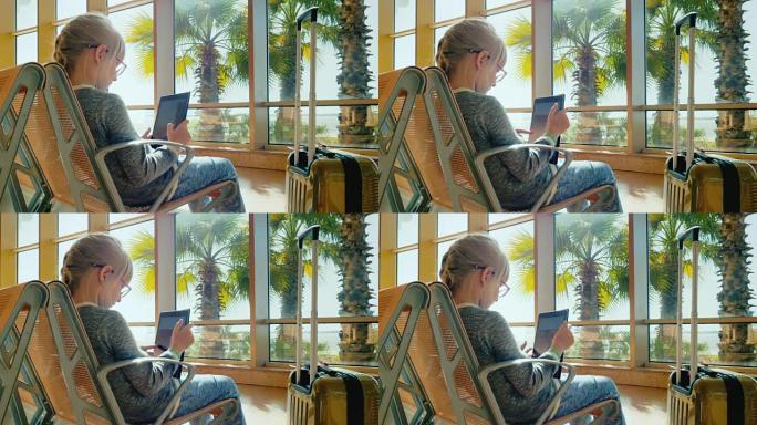 戴眼镜的金发女郎正在平板电脑上玩。坐在机场航站楼靠近阳光普照和棕榈树生长的窗户。休假回家