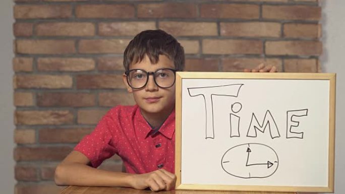 孩子坐在办公桌前拿着挂图，背景红砖墙上刻字时间