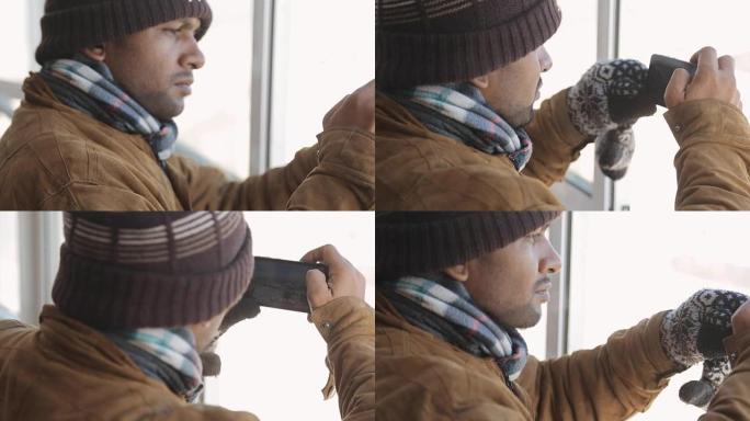 严肃的穿着暖和的男人-印度教徒在冬天通过窗户用智能手机拍照