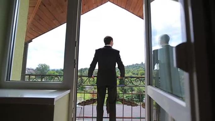 穿着黑色西装的英俊自信的新郎站在阳台上，靠在栏杆上，看着背景上的绿色森林。后视图