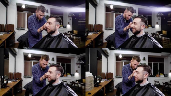 在barbeshop中，时尚微笑的大胡子男人坐在黑色的peignoir上。理发师穿着便服站在顾客身后