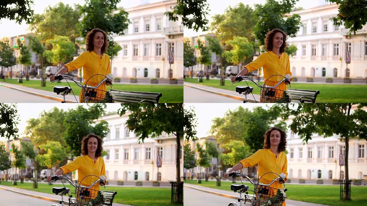清晨，穿着黄色长裙的微笑迷人的女人拿着城市自行车的车把，篮子里放着鲜花。快乐的女人享受她的假期