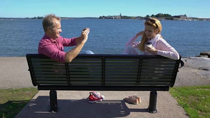 一对在海边的长凳上。年轻人正在用智能手机给她的女朋友拍照。