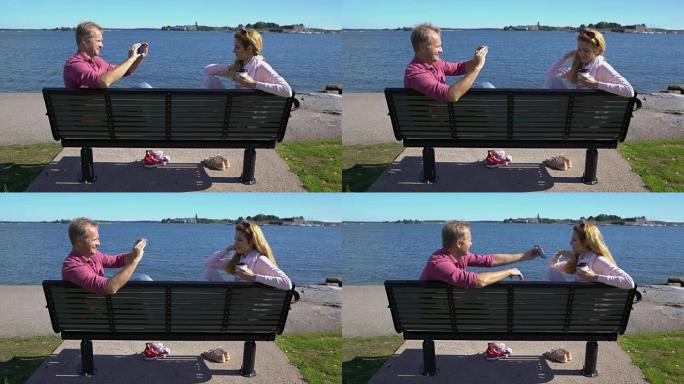 一对在海边的长凳上。年轻人正在用智能手机给她的女朋友拍照。