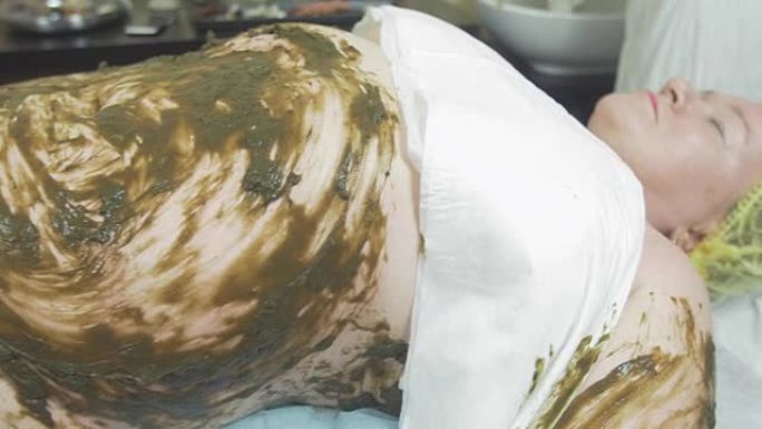 成年胖女人躺在肚子上，左手躺在海藻混合物中。水疗包装