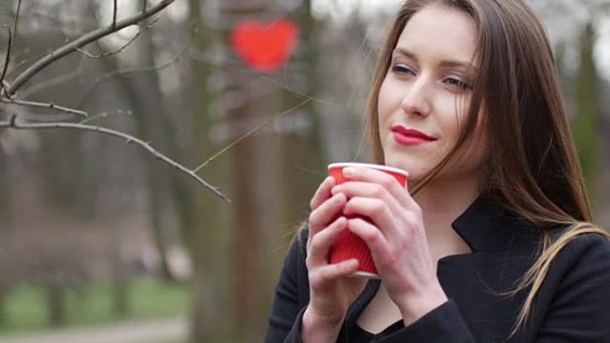 年轻美丽的金发时髦女人的肖像在秋天的公园背景上摆姿势，喝一杯外卖咖啡