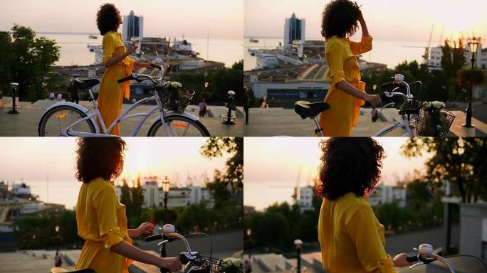 黎明时分，穿着黄色长裙的迷人黑发女人站在她的城市自行车旁边，拿着车把，篮子里放着鲜花，享受清晨。镜头