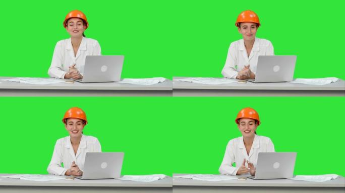 微笑的女工程师对着坐在桌子上的相机说话，绿色屏幕上有很多纸，色度键