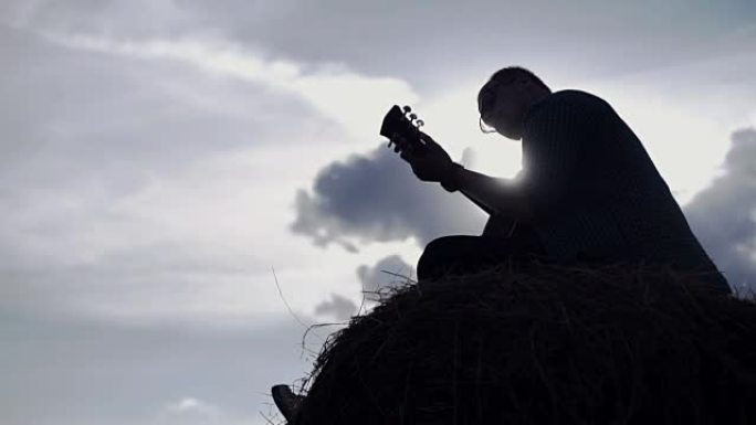 穿着方格衬衫的家伙坐在干草堆上的田野里弹吉他