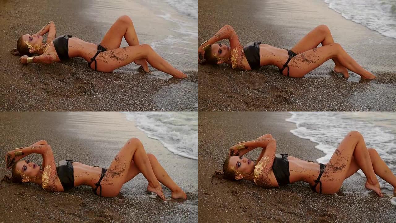 穿着黑色皮革比基尼的性感苗条模特躺在海浪中的海滩上。诱人的年轻女子，专业的艺术金色化妆和海边沙滩上的