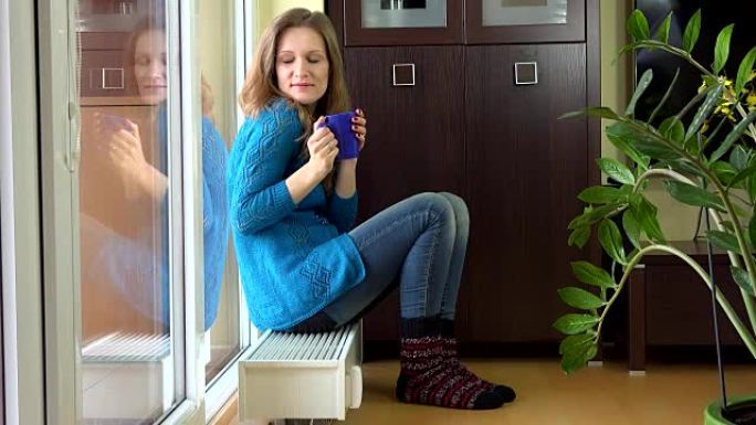 穿着暖和衣服的幸福女人喝茶，梦想坐在温暖的散热器上。
