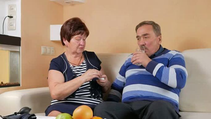 一位年长的妇女在家中沙发上给丈夫喝了一杯水。穷人的幸福。照顾妻子的丈夫