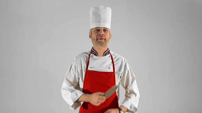 英俊的成年厨师转身，他用刀交叉双臂，在灰色背景上点头