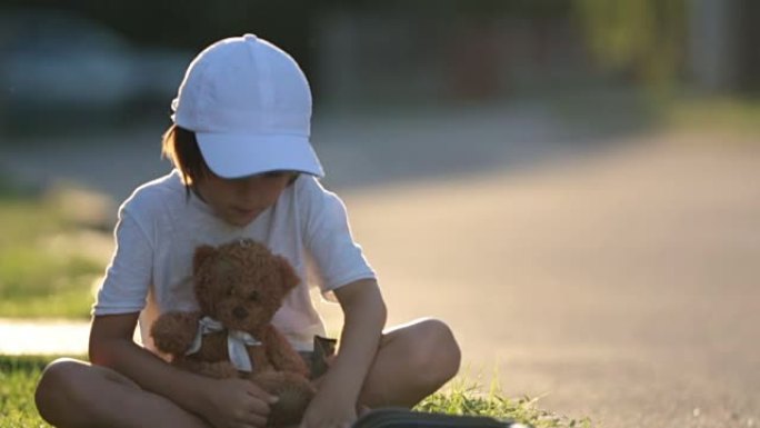 漂亮的小男孩，在街上看书，和泰迪熊坐在一起，美丽的日落之光
