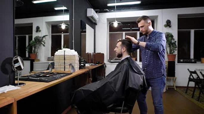 穿着便服的年轻理发师在剪头发前站着梳理客户。微笑的男人坐在椅子上，上面覆盖着黑色的peignoir，