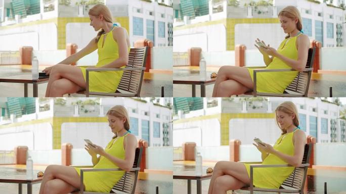 穿着黄色连衣裙的微笑幸福女人坐在游泳池附近的椅子上。使用她的智能手机