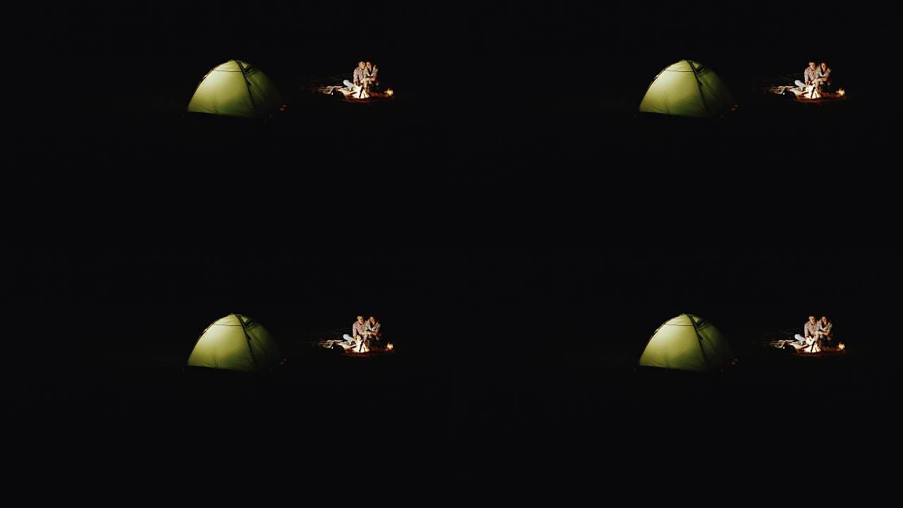 浪漫的情侣晚上在篝火旁放松，站在帐篷旁边，灯光照耀着