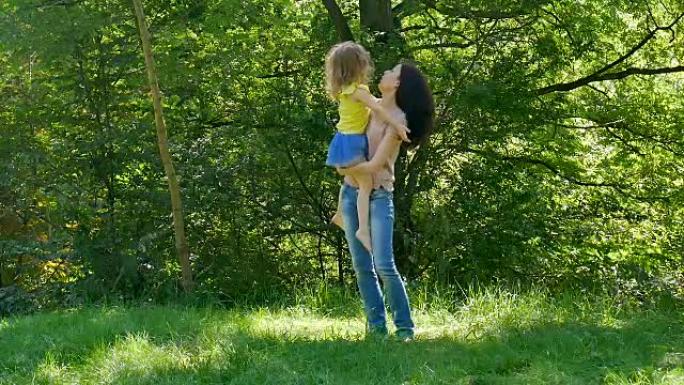 在阳光明媚的温暖日子里，幸福的家庭在公园里共度时光。黑发母亲抱起女儿旋转。小女孩亲吻妈妈。