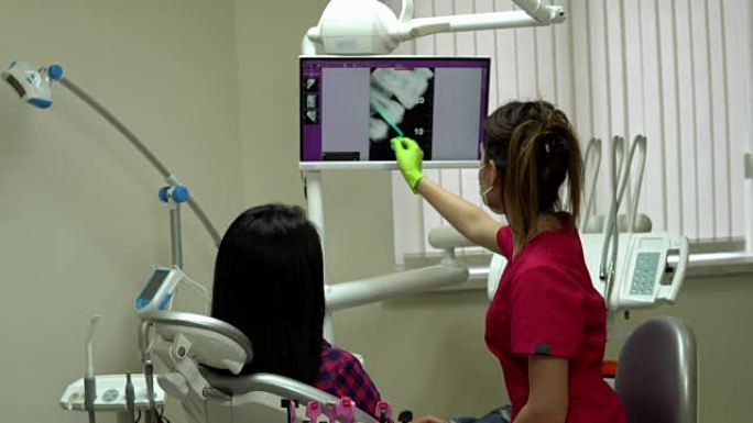 年轻的女医生用x光指着屏幕，解释她的病人需要治疗。年轻的女人坐在牙医的椅子上，看着监视器。4k拍摄