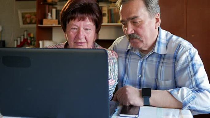 一对老年夫妇正坐在家里的笔记本电脑旁。一个女人读新闻，一个留着小胡子的男人坐在他旁边说话