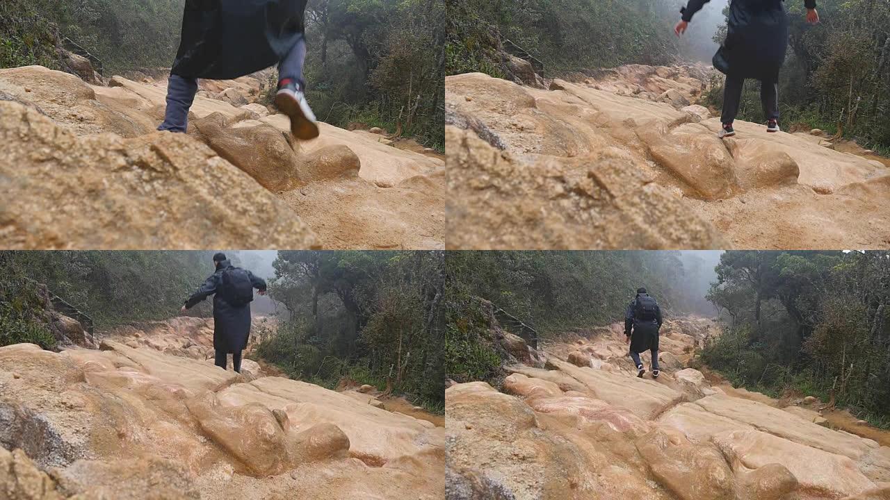 穿着雨衣的年轻人在旅行中在岩石小径上跳跃和慢跑。徒步旅行的人，背包在潮湿的森林里的石头地上奔跑。男性