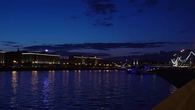 蓝色小时。河上的夜市灯光。海滨城市公园的景色。光线明亮的小船飘过。行人走在傍晚的长廊上。夜间汽车交通