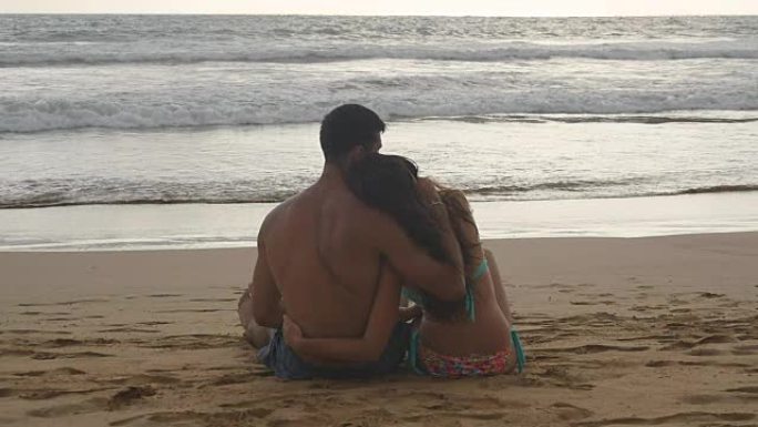 年轻浪漫的夫妇正坐在沙滩上拥抱着美丽的日落。一个女人和一个男人一起坐在海边的沙滩上，欣赏海洋和风景。