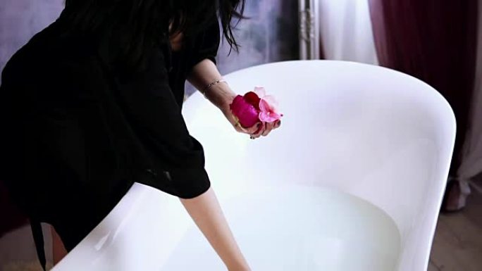 穿着黑色浴袍的年轻迷人女人向装满牛奶的浴缸扔花，准备洗个热水澡。慢动作镜头