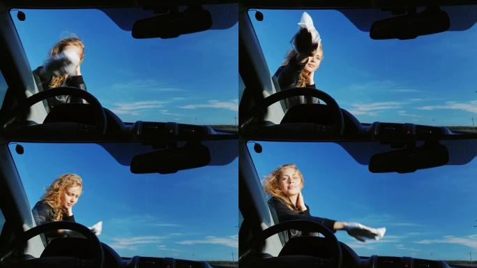 年轻迷人的女人在擦汽车的挡风玻璃。从车里看。在晴朗的日子里，以红蓝的天空为背景