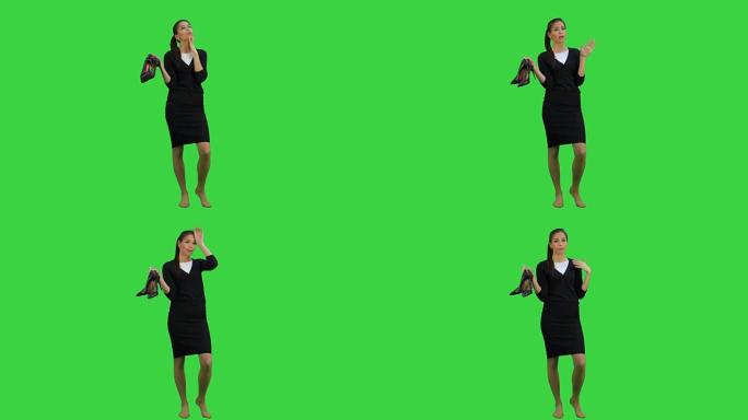 疲惫的年轻女商人赤脚站着，在绿色屏幕上拿着高跟鞋，色键