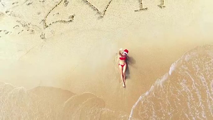鸟瞰图。戴着圣诞老人帽子的年轻女子躺在海滩上。圣诞假期。沙子上的2017铭文